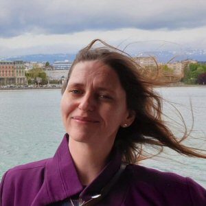 A person with long hair smiles, standing near a body of water on a windy day, as buildings and a stunning drone show light up the background.