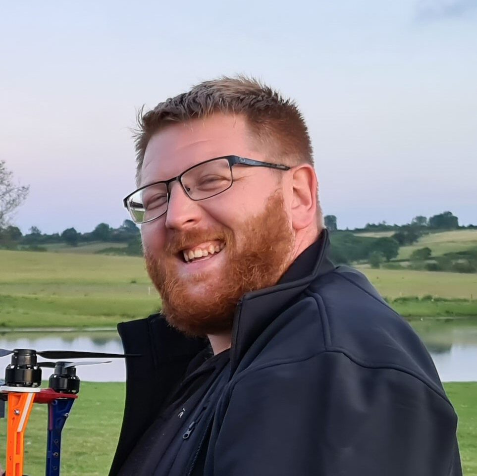 A smiling man with glasses and a beard, clad in a black jacket, stands outdoors near a tranquil body of water and green fields, as a mesmerizing drone show lights up the sky above.