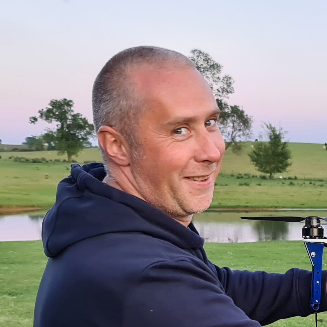 Smiling man with a shaved head wearing a dark hoodie, standing outdoors near a pond and grassy field with trees, as the sky above hints at an upcoming drone show.